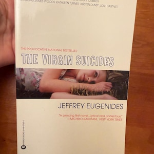 Op de afbeelding: Een paperback boek met de titel "THE VIRGIN SUICIDES" van Jeffrey Eugenides. De omslag toont een persoon die in het gras ligt. Het boek is een "provocerende nationale bestseller" en is nu een grote film.