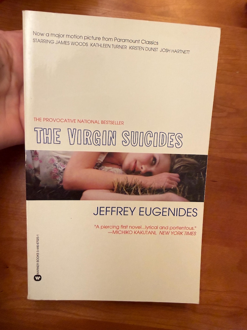 Op de afbeelding: Een paperback boek met de titel "THE VIRGIN SUICIDES" van Jeffrey Eugenides. De omslag toont een persoon die in het gras ligt. Het boek is een "provocerende nationale bestseller" en is nu een grote film.