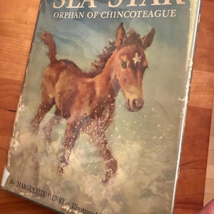 Puede incluir: Portada de libro vintage titulada "SEA STAR Orphan of Chincoteague". La portada presenta un caballo marrón corriendo por el agua. El título está en letras azules grandes y en negrita. El nombre del autor, Marguerite Henry, está en la parte inferior.