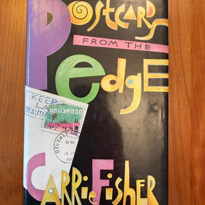 May include: A hardcover book titled "Postcards from the Edge" by Carrie Fisher. The cover features a black background with colorful text and a purple "P" and a green "e". A postcard graphic is on the lower left.