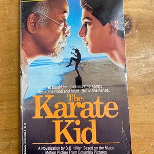 Puede incluir: Portada del libro The Karate Kid. La portada presenta a dos hombres enfrentados con una figura de karate en el medio. El título es naranja, con el texto "Le enseñó el secreto del Karate reside en la mente y el corazón. No en las manos."