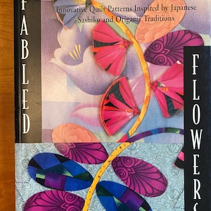 May include: A book titled "Fabled Flowers" by Kumiko Sudo. The book cover features a colourful quilt design inspired by Japanese Sashiko and Origami traditions. The quilt design depicts a stylised flower with pink, purple, and blue fabric pieces.