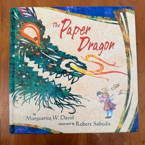 May include: A hardcover children's book titled "The Paper Dragon." The cover features a green and blue dragon's head with orange flames, a child, and the names of the author and illustrator.