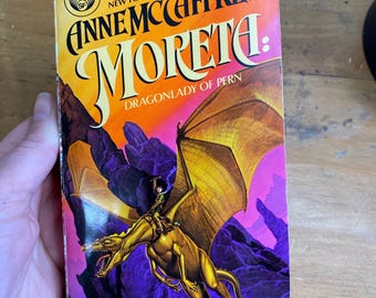 May include: A paperback book cover with a yellow dragon flying in front of a purple and pink background. The title is "Moreta: Dragonlady of Pern" by Anne McCaffrey.