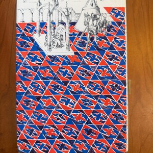 May include: Book cover with a geometric pattern in red, white, and blue. The top features a black and white illustration of a medieval scene with figures and architecture. A tan bookmark is attached.