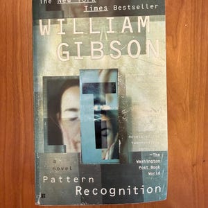 Puede incluir: Un libro de tapa dura titulado "Pattern Recognition" de William Gibson. La portada presenta una imagen borrosa de un rostro, con el título y el nombre del autor en letras grandes y en negrita. El libro es un éxito de ventas del New York Times.