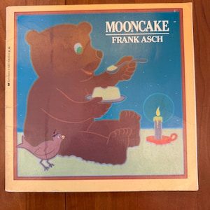 May include: A children's book cover featuring a brown bear holding a spoon and a plate with a yellow dessert. The title "MOONCAKE" and author "FRANK ASCH" are displayed in white text against a blue starry background. A small bird and a candle are also present.