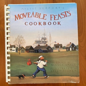 Könnte beinhalten: Ein Kochbuch mit dem Titel "Mystic Seaport's Moveable Feasts Cookbook" mit einer Illustration eines Seemanns, der eine Tasche auf einem Stock trägt, und einem Hund, der neben ihm geht. Der Hintergrund zeigt eine Hafenszene mit einem Schiff und Häusern.