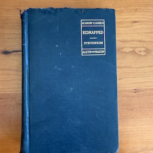 May include: A dark blue hardcover book titled "Kidnapped" by Stevenson. The book is part of the "Academy Classics" series and published by Allyn and Bacon. The book is on a wooden surface.