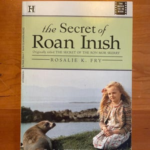 May include: A paperback book titled The Secret of Roan Inish by Rosalie K. Fry. The cover features a young person with long blonde hair sitting on grass next to a seal. The text includes the movie tie-in label and the price of $5.95 in Canada.