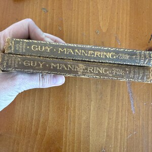 May include: Two antique books titled "Guy Mannering" by Walter Scott. The spines are brown with gold lettering, and the volumes are labeled "Vol. One" and "Vol. Two." The books are held by a hand, with a wooden surface in the background.