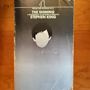 Könnte beinhalten: Ein Taschenbuch mit dem Titel The Shining von Stephen King mit einem schwarz-blauen Farbverlauf auf dem Cover. Das Cover zeigt eine stilisierte Illustration eines Kinderkopfes und den Text FROM THE AUTHOR OF IT, THE SHINING, A MASTERPIECE OF MODERN HORROR.