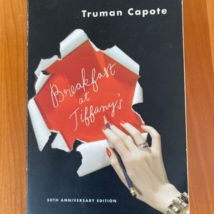May include: A black and white book cover with the title "Breakfast at Tiffany's" in red script. The author, Truman Capote, is listed above the title. The cover features a hand holding a pencil and a piece of paper with the title written on it. The book is a 50th anniversary edition.
