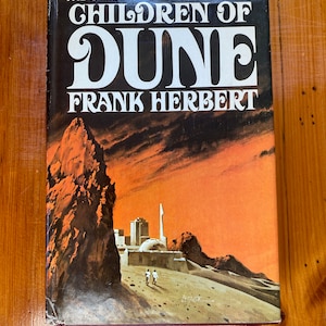 May include: A vintage paperback book titled "Children of Dune" by Frank Herbert. The cover features a desert landscape with a city and two figures, set against an orange and brown sky. The title is in large white letters.