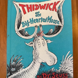 Könnte beinhalten: Ein Kinderbuch mit festem Einband mit dem Titel Thidwick The Big-Hearted Moose von Dr. Seuss. Das Cover zeigt eine Illustration eines weißen Elchs auf hellblauem Grund, der Titel steht in Rot und Blau auf einem Banner zwischen dem Geweih.