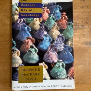 May include: Book cover for "The Redneck Way of Knowledge" by Blanche McCrary Boyd. The cover features a collection of colorful, doll-like figures. The title is in a blue and gold rectangular box. The author's name is in a gold box.