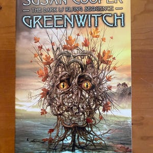 Pode incluir: Um livro de bolso intitulado Greenwitch de Susan Cooper, parte da série The Dark Is Rising Sequence. A capa apresenta uma figura central feita de galhos e ramos castanhos entrelaçados com olhos amarelos brilhantes e folhas de outono.