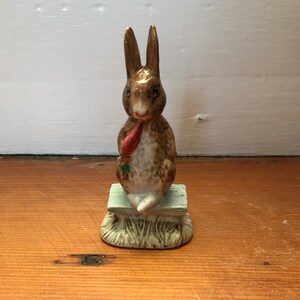 May include: A brown ceramic figurine of a rabbit sitting on a green bench with a red carrot in its mouth. The rabbit is sitting on a base that looks like grass.