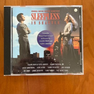 Könnte beinhalten: Eine CD-Hülle für den Soundtrack des Films "Schlaflos in Seattle". Das Cover zeigt zwei Personen, die nach oben schauen, mit einer Skyline im Hintergrund. Die CD-Hülle enthält die Namen der Künstler und Songs.