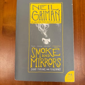 Könnte beinhalten: Ein graues Taschenbuch mit dem Titel "Smoke and Mirrors" von Neil Gaiman. Das Cover zeigt gelben Text und eine Rauchillustration. Das Buch ist eine Sammlung von Kurzgeschichten und Illusionen.
