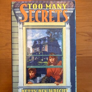 Peut inclure: Couverture du livre "Too Many Secrets" de Betty Ren Wright. L'illustration montre une fenêtre avec deux enfants, un chien et une maison. Le titre est en orange, le nom de l'auteur en bas.
