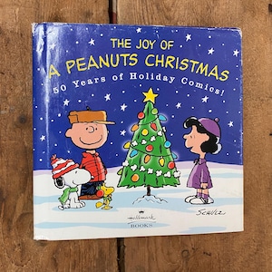 May include: A blue book cover with the title "The Joy of A Peanuts Christmas" and the subtitle "50 Years of Holiday Comics!"  The cover features Charlie Brown, Snoopy, Lucy, and a Christmas tree with ornaments and lights. The book is published by Hallmark Books.