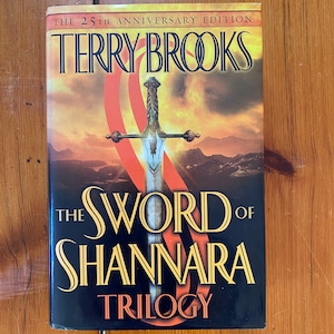 Puede incluir: La edición del 25 aniversario de La Espada de Shannara de Terry Brooks. La portada del libro presenta una espada sobre un fondo rojo y naranja.