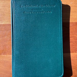 May include: A teal green hardcover book titled "The Manhood of the Master" by Harry Emerson Fosdick. The title and author's name are embossed on the cover. A white ribbon bookmark is visible.