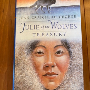 Puede incluir: Una portada de libro con fondo azul y una capucha de piel blanca. El título es "Julie of the Wolves Treasury" de Jean Craighead George. Un lobo camina en el fondo.