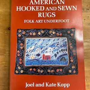 May include: A red book titled "American Hooked and Sewn Rugs: Folk Art Underfoot" by Joel and Kate Kopp. The cover features a colorful hooked rug depicting a landscape with a house, trees, and clouds.