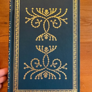 May include: A dark blue hardcover book featuring intricate gold foil stamping on the front cover. The design includes a rectangular border and two symmetrical floral motifs composed of delicate vines and small star-shaped elements.
