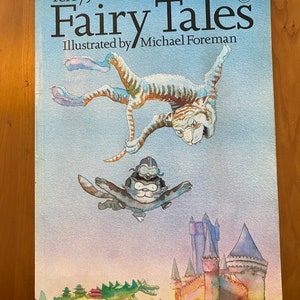 Puede incluir: Un libro titulado "Fairy Tales" de Terry Jones, ilustrado por Michael Foreman. La portada presenta una ilustración de acuarela de un tigre volando por el aire con dos gatos más pequeños que lo montan. Un castillo y un dragón verde son visibles en el fondo.