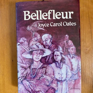 Op de afbeelding: Een boekomslag met een paarse achtergrond en de titel "Bellefleur" van Joyce Carol Oates. De omslag toont een schilderij van een groep mensen, waaronder een vrouw die een kat vasthoudt.