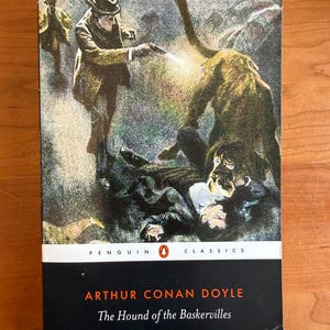 May include: A Penguin Classics edition of Arthur Conan Doyle's "The Hound of the Baskervilles." The cover art depicts a dramatic scene with figures, a hound, and a gun firing. The title and author's name are in white and orange on a black background.