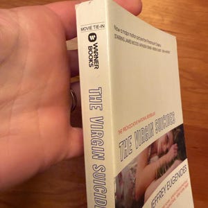 Op de afbeelding: Een paperback boek getiteld "The Virgin Suicides" van Jeffrey Eugenides. De omslag toont een foto en tekst. De rug leest "The Virgin Suicides" en "Warner Books". Het is een film-tie-in editie.