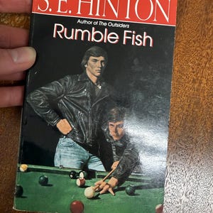 May include: A paperback book titled "Rumble Fish" by S.E. Hinton. The cover features two men in leather jackets, one standing and one playing pool. The background is black with a green pool table.