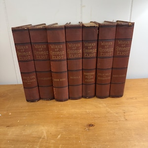 Puede incluir: Una colección de libros antiguos con cubiertas granates. Las lomos muestran el título "Obras de George Eliot" en letras doradas. Los títulos incluyen "Adam Bede", "Romola" y "Poemas y Ensayos". Los libros están colocados sobre una superficie de madera.