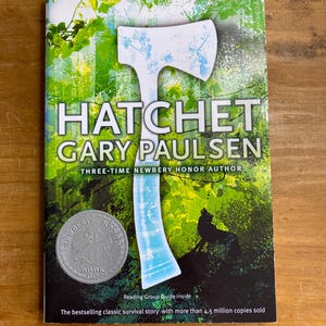 Puede incluir: Un libro de bolsillo titulado "Hatchet" de Gary Paulsen. La portada presenta un hacha blanca sobre un fondo verde y azul. Un sello de honor Newbery plateado está en la parte inferior izquierda. El texto dice "La historia de supervivencia clásica más vendida con más de 4,5 millones de copias vendidas."