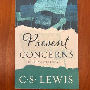May include: A book titled "Present Concerns" by C.S. Lewis. The cover features a blue and white illustration of flags and signs. The title is in a decorative font, with "Journalistic Essays" below. The book is on a wooden surface.