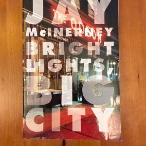May include: A book cover for "Bright Lights, Big City" by Jay McInerney. The title is in large, transparent white letters over a nighttime street scene with red neon signs. The author's name is at the top.