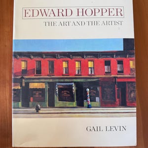 Puede incluir: Un libro titulado "Edward Hopper: The Art and the Artist" de Gail Levin. La portada presenta una pintura de un edificio de ladrillo rojo con tiendas y un cielo azul. El libro está sobre una superficie de madera.