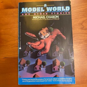 May include: A book cover with a black background and a blue bottom border. The title is "A Model World and Other Stories" by Michael Chabon. The cover features a hand holding a small model house with a tree in front of it. The background shows a series of model houses.