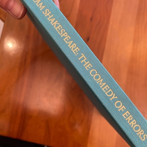 May include: A teal book spine with gold lettering. The text reads "WILLIAM SHAKESPEARE: THE COMEDY OF ERRORS". The book appears to be hardcover and is set against a wooden surface.