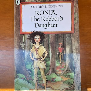 Può includere: Copertina del libro con il titolo "RONIA, The Robber's Daughter" di Astrid Lindgren. L'illustrazione raffigura un giovane in una foresta, con un'altra figura sullo sfondo. La copertina ha un bordo marrone e uno sfondo marrone chiaro.