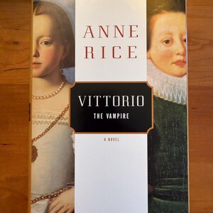 以下が含まれることがあります： アン・ライス著『Vittorio the Vampire』のハードカバー本。表紙は分割されたパネルデザインで、左側に白いドレスを着た人物、右側に緑色の衣服を着た人物の肖像画が描かれており、中央の白い帯で分けられています。