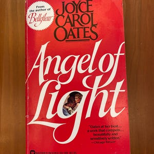Könnte beinhalten: Ein rotes Taschenbuch mit dem Titel "Angel of Light" von Joyce Carol Oates. Das Cover hat weiße Schrift und ein Foto von zwei Personen. Herausgegeben von Warner Books.