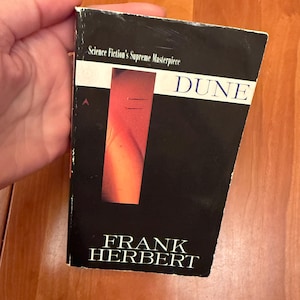May include: A paperback book titled "Dune" by Frank Herbert. The cover is predominantly black with a vertical orange and red design. The title is in white, and the text "Science Fiction's Supreme Masterpiece" is at the top.
