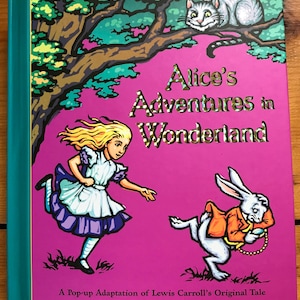 May include: A hardcover book titled Alice's Adventures in Wonderland, a pop-up adaptation by Robert Sabuda. The cover features a bright pink background with a blonde character in a blue dress, a white rabbit in an orange coat, and a cat in a tree.