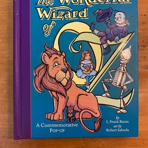 May include: A hardcover book titled The Wonderful Wizard of Oz, A Commemorative Pop-Up by L. Frank Baum with art by Robert Sabuda. The blue cover features illustrations of a lion, a girl, a scarecrow, and a tin man against a large yellow letter O.