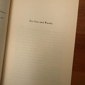 Op de afbeelding: Open boek met de tekst "For Gus and Wanda" in zwarte letters op een cr&egrave;mekleurige pagina. Het boek ligt op een houten oppervlak.